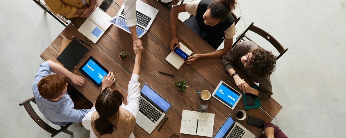 A diverse team of professionals collaborating around a wooden conference table with laptops, tablets, notebooks, and coffee cups—two people shaking hands in agreement during a productive business meeting