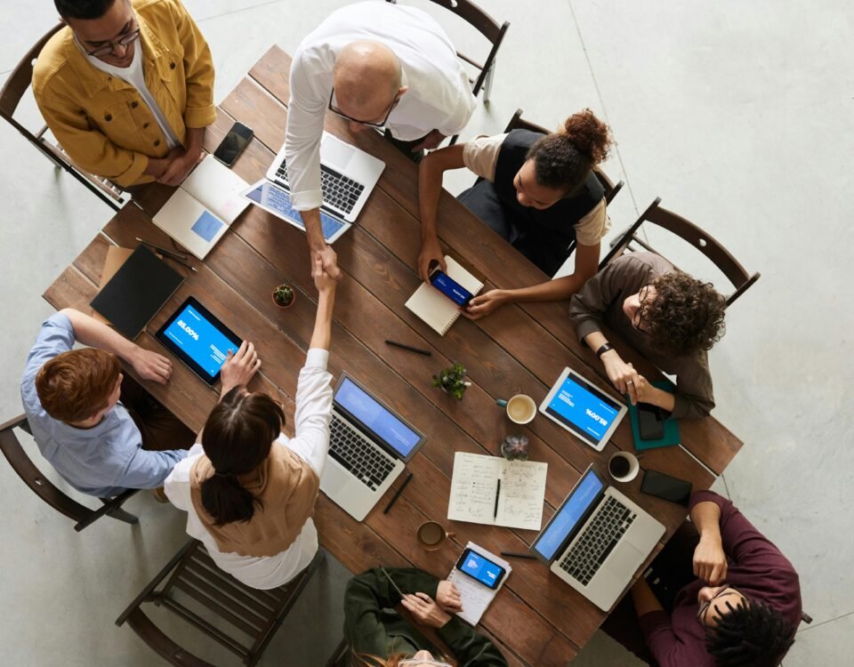 A diverse team of professionals collaborating around a wooden conference table with laptops, tablets, notebooks, and coffee cups—two people shaking hands in agreement during a productive business meeting